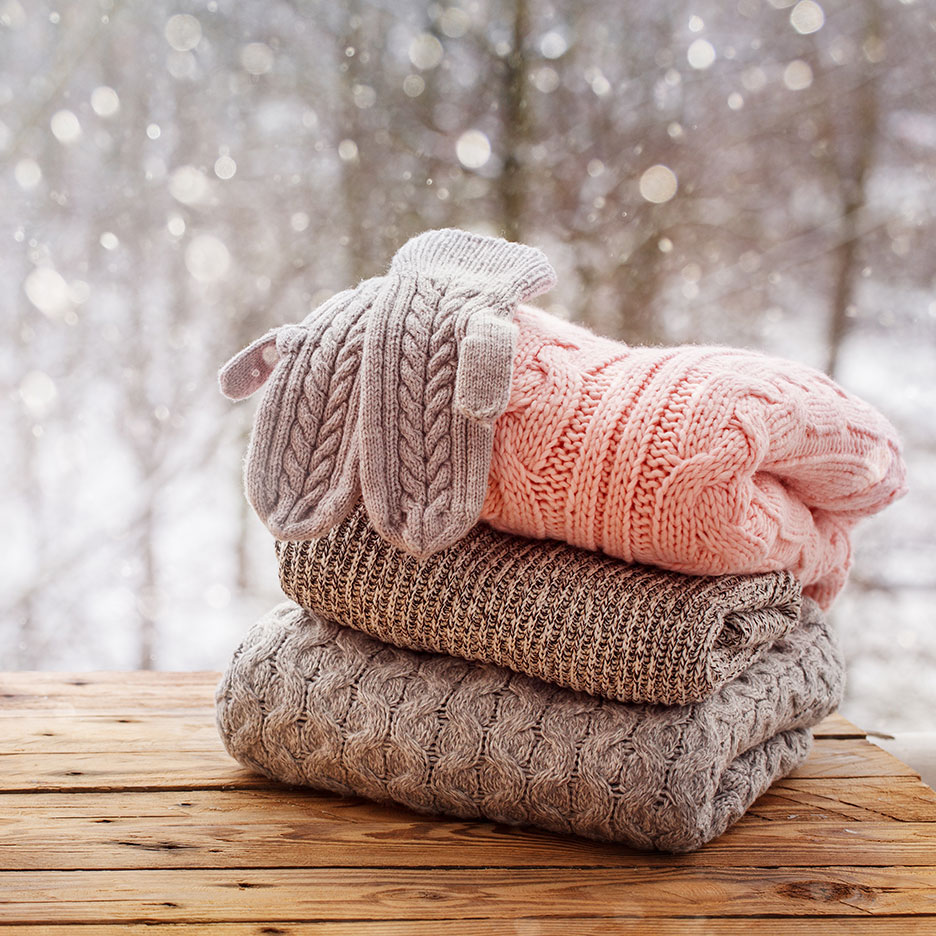 Knitted sweaters and mittens on a wooden table with a snowy background.