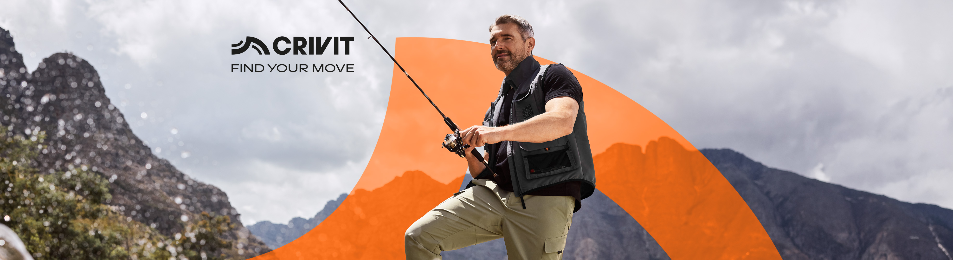 Man in a fishing vest and cargo pants holding a fishing rod, with mountains in the background.