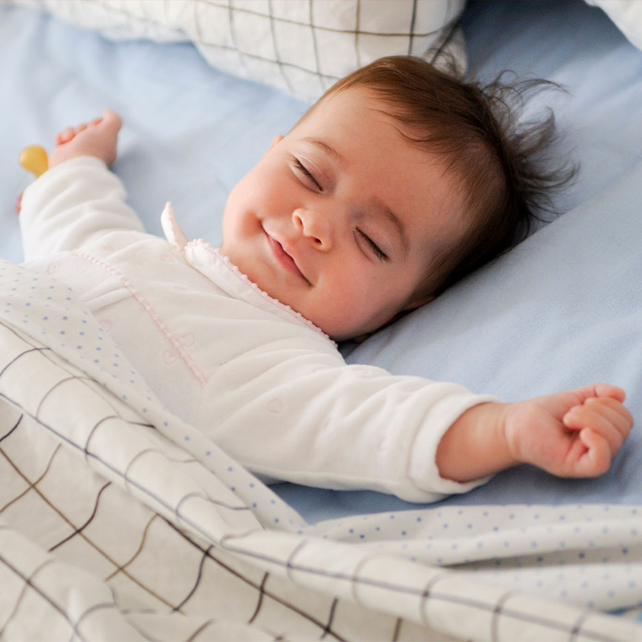 A sleeping baby in a white sleepsuit, covered by a patterned duvet.
