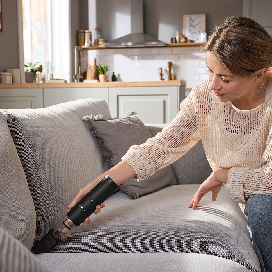 Woman vacuuming a sofa with a handheld vacuum cleaner in a living room.
