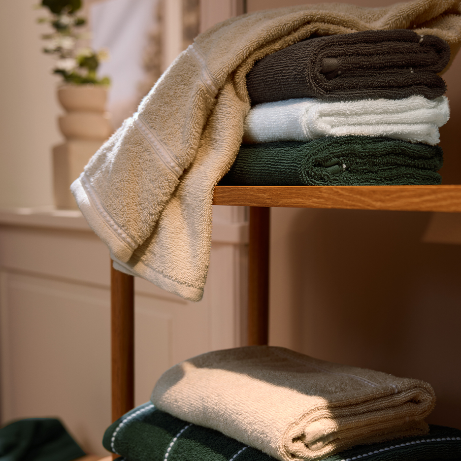Stacked beige, dark brown, white, and dark green towels on a wooden shelf.