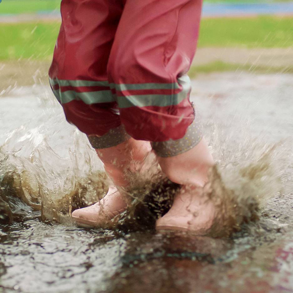 Child in red waterproof trousers and boots jumping in a puddle.