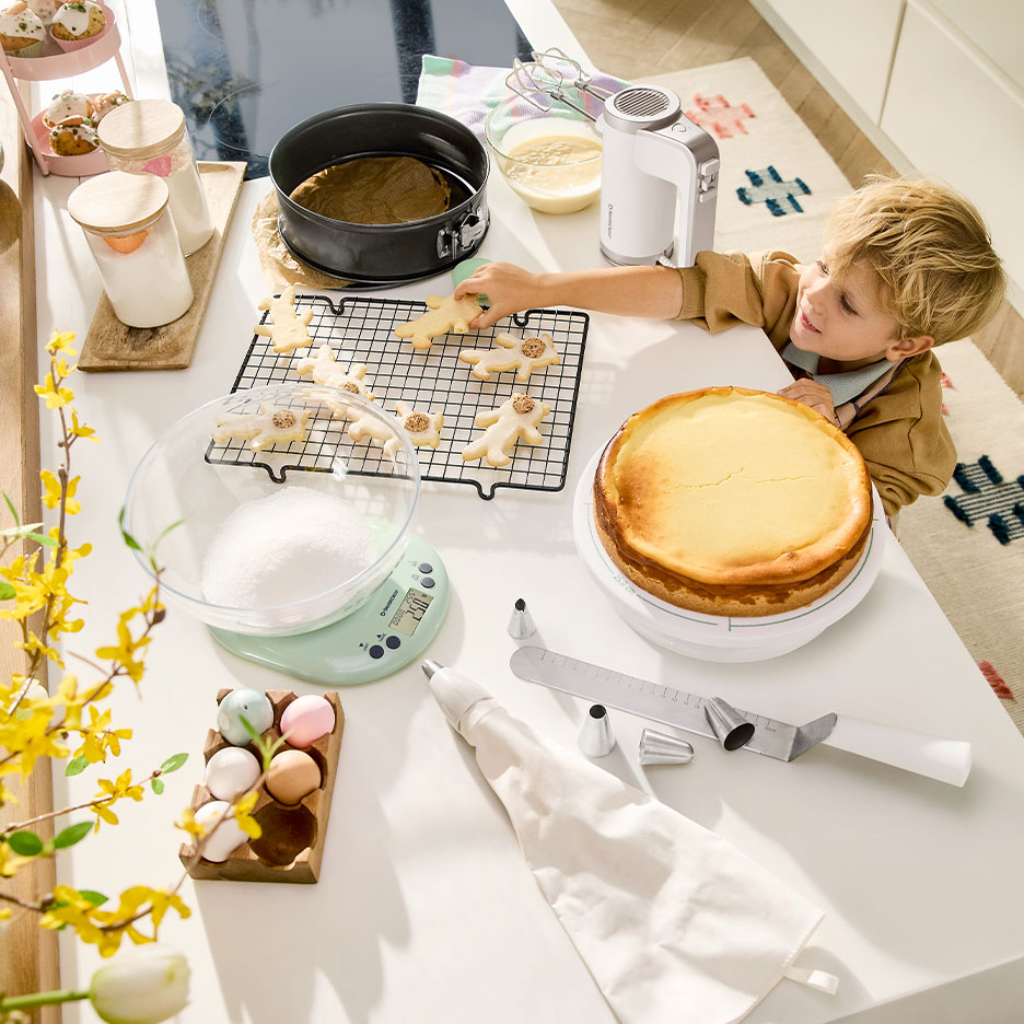 A young boy decorates biscuits, with a cheesecake and baking utensils on the table.