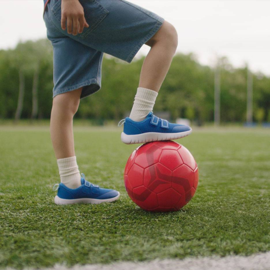 Child in shorts and blue sneakers, with one foot on a red soccer ball.