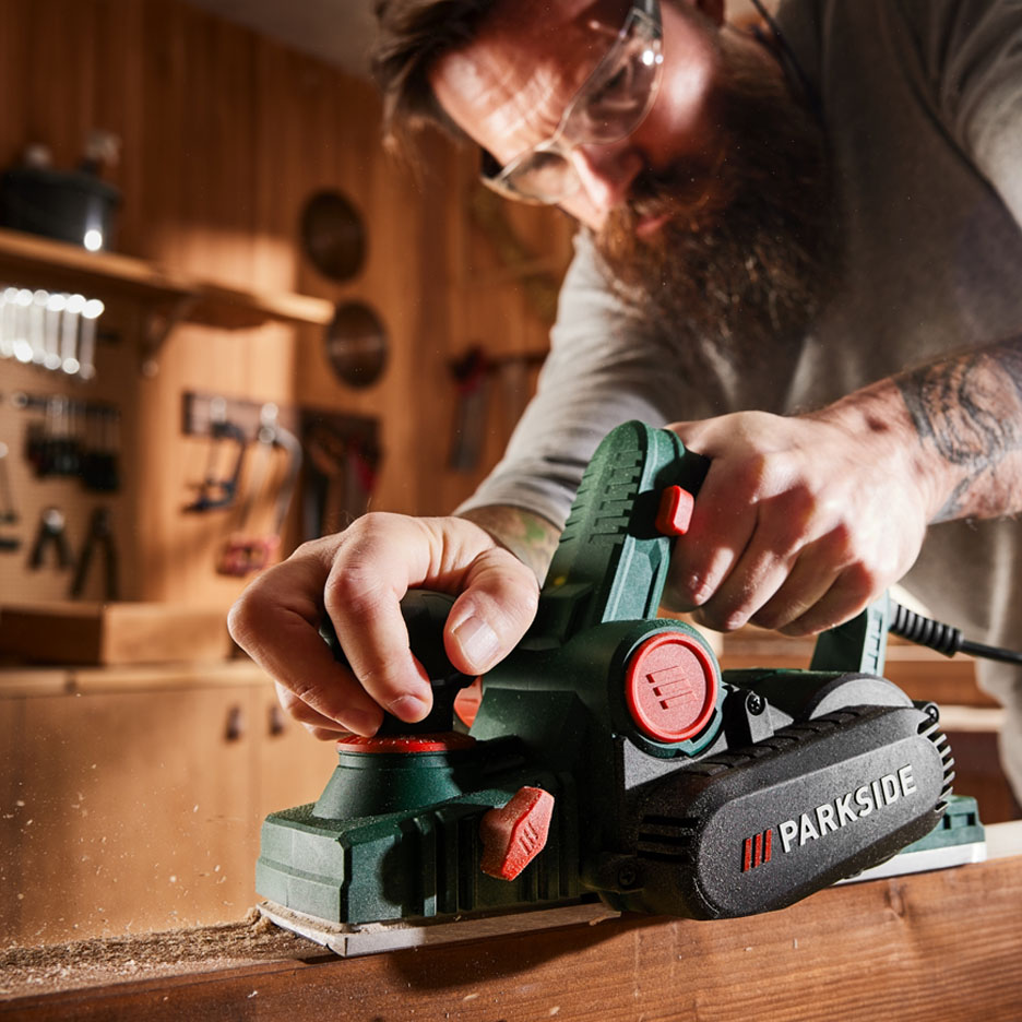 Man in safety glasses using a PARKSIDE electric planer on wood in a workshop.