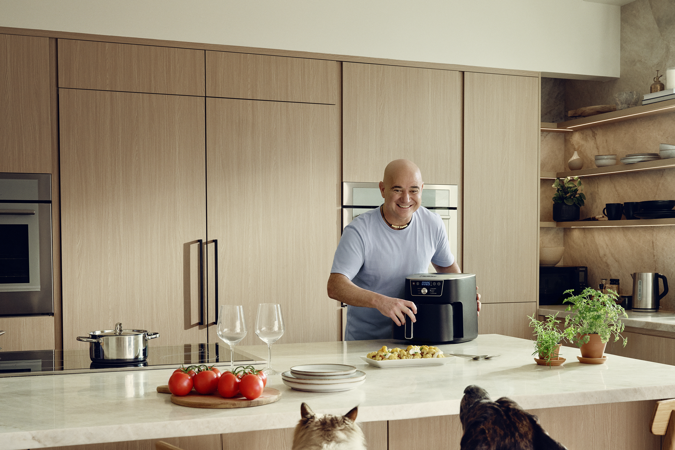 Andre Agassi in a kitchen with an air fryer, preparing food while dogs watch.