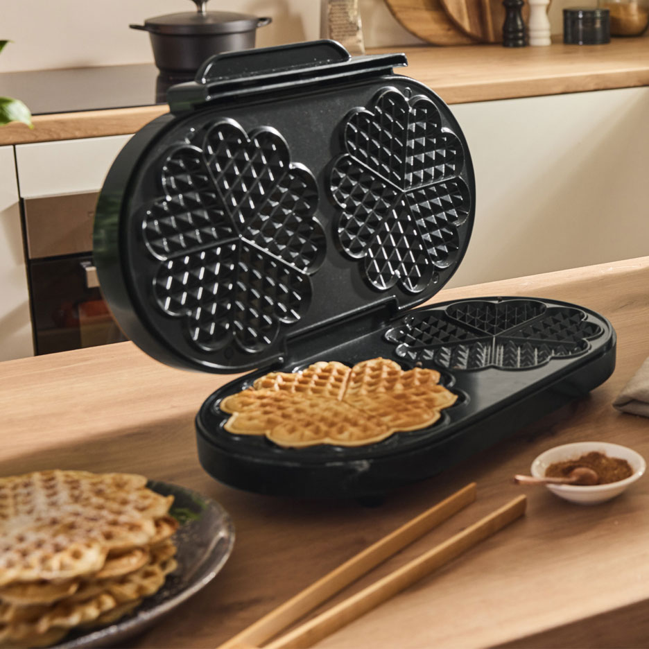 Open waffle maker with a heart-shaped waffle on a wooden table.