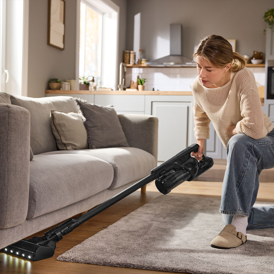 Woman cleaning living room with a cordless stick vacuum cleaner under the sofa.