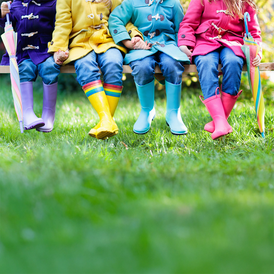 Children in colorful raincoats and wellington boots sitting on a bench with umbrellas.