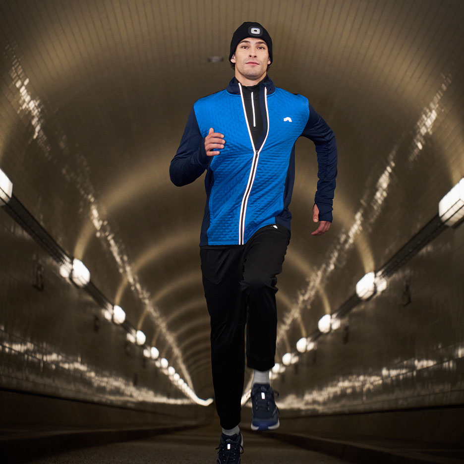 Man in running jacket, pants, and beanie with headlamp running in a tunnel.
