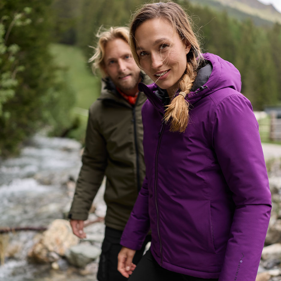Two hikers in winter jackets by a river in the mountains.
