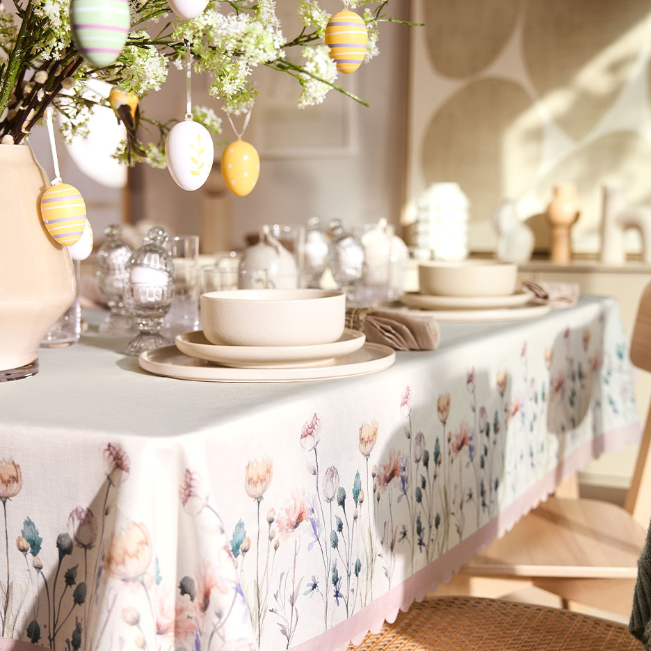 Easter table with a floral tablecloth, decorated with hanging eggs and dinnerware in neutral tones.