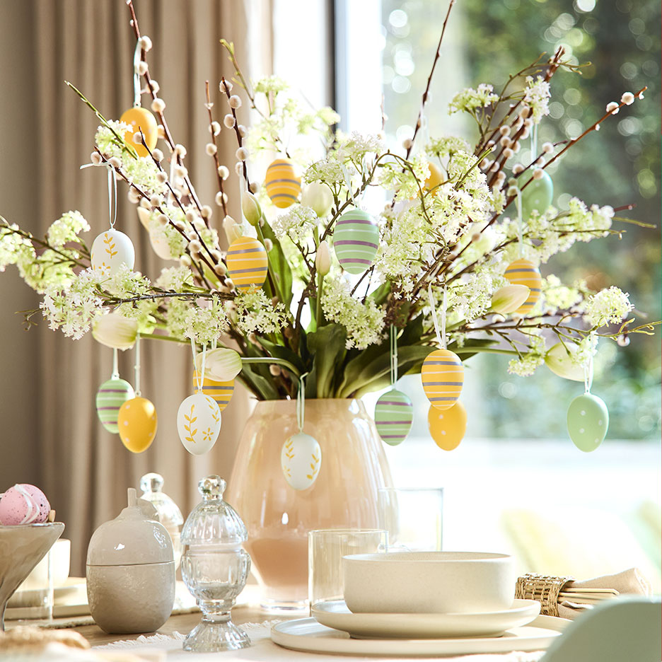 Vase with willow branches, white flowers, and colorful Easter eggs on a set table.