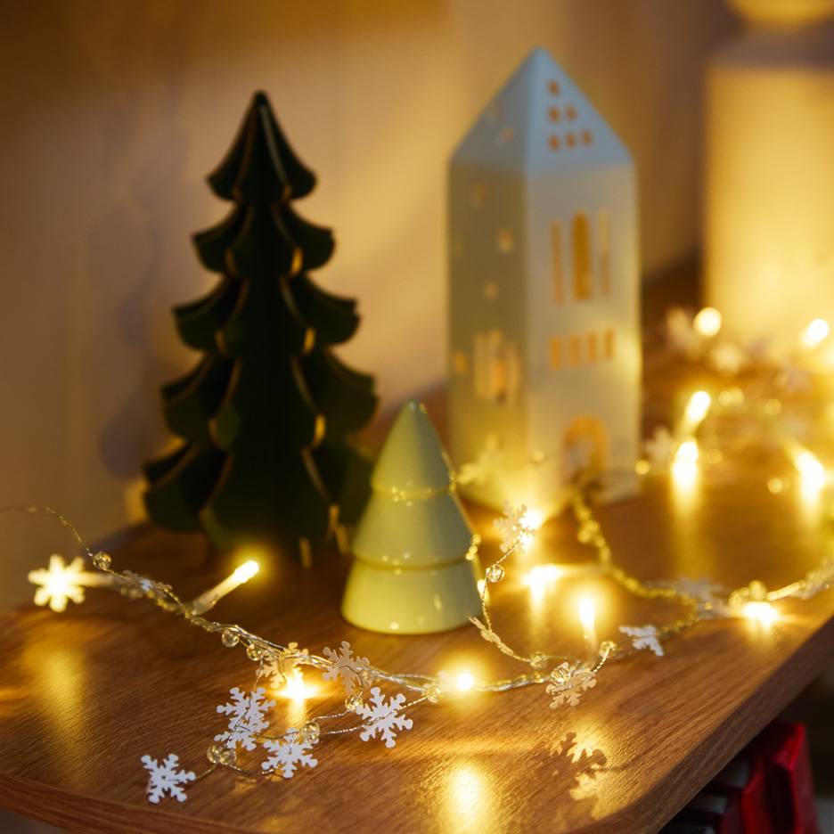 Christmas decorations: a dark green tree, a light green tree, a white house, and a snowflake string light.