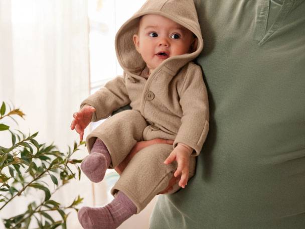 Baby in a beige hooded romper, held by an adult in a green t-shirt.