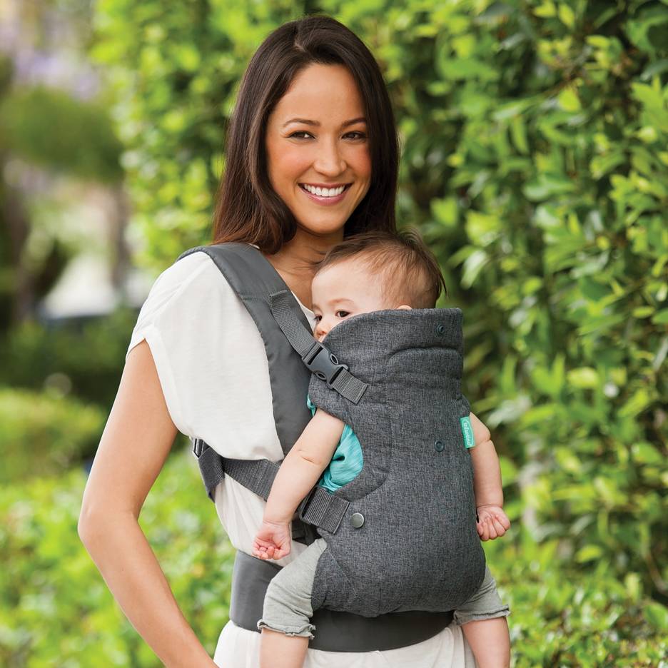 Smiling woman in a grey baby carrier holding a baby outdoors.
