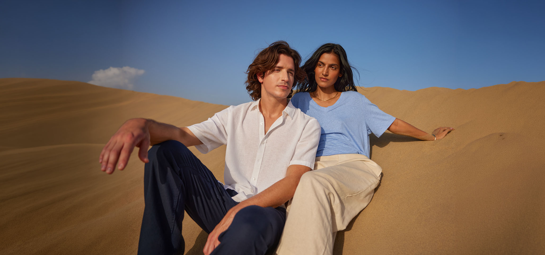 Man and woman sitting on sand dunes, wearing linen shirts and trousers.