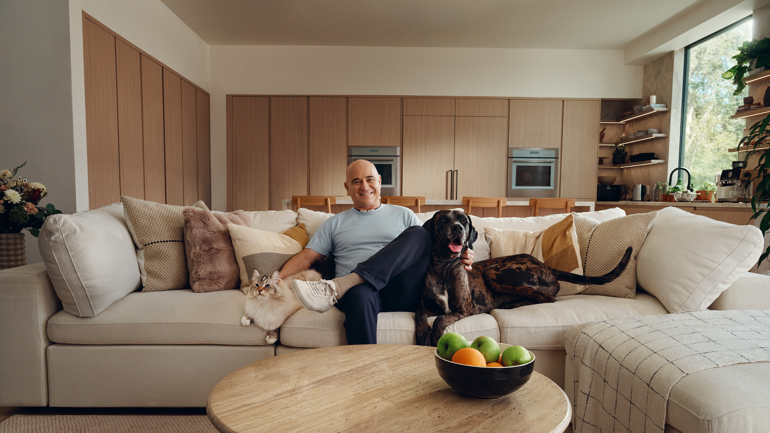 Andre Agassi sits on a sofa with a cat and dog, with a fruit bowl on the coffee table.